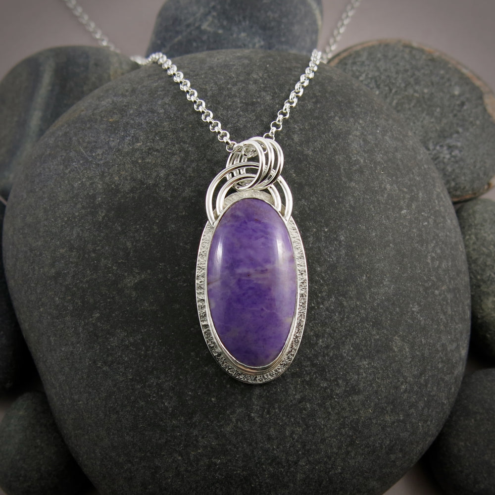 A sterling silver necklace with a purple jade gemstone is displayed on a black beach rock.  The stone is slightly mottled and there is a subtly textured silver "halo" surrounding the gem.  There is also a double arc of wire atop the pendant.
