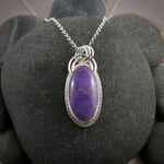 A sterling silver necklace with a purple jade gemstone is displayed on a black beach rock.  The stone is slightly mottled and there is a subtly textured silver "halo" surrounding the gem.  There is also a double arc of wire atop the pendant.
