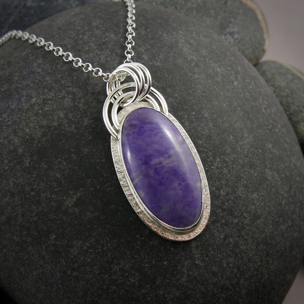 A sterling silver necklace with a purple jade gemstone is displayed on a black beach rock.  The stone is slightly mottled and there is a subtly textured silver "halo" surrounding the gem.  There is also a double arc of wire atop the pendant.