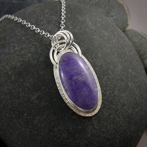 A sterling silver necklace with a purple jade gemstone is displayed on a black beach rock.  The stone is slightly mottled and there is a subtly textured silver "halo" surrounding the gem.  There is also a double arc of wire atop the pendant.