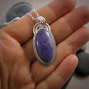 A sterling silver necklace with a purple jade gemstone is displayed on a hand.  The stone is slightly mottled and there is a subtly textured silver "halo" surrounding the gem.  There is also a double arc of wire atop the pendant.