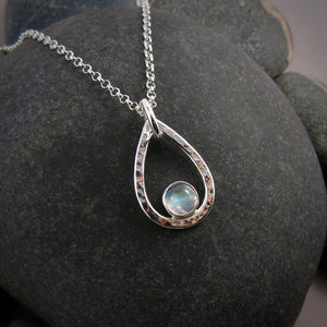 A teardrop shaped necklace is displayed on a black beach rock.  The hammer textured drop is accented by a round rainbow moonstone that sits in the curve at the bottom of the pendant.  A silver rolo chain completes the piece.