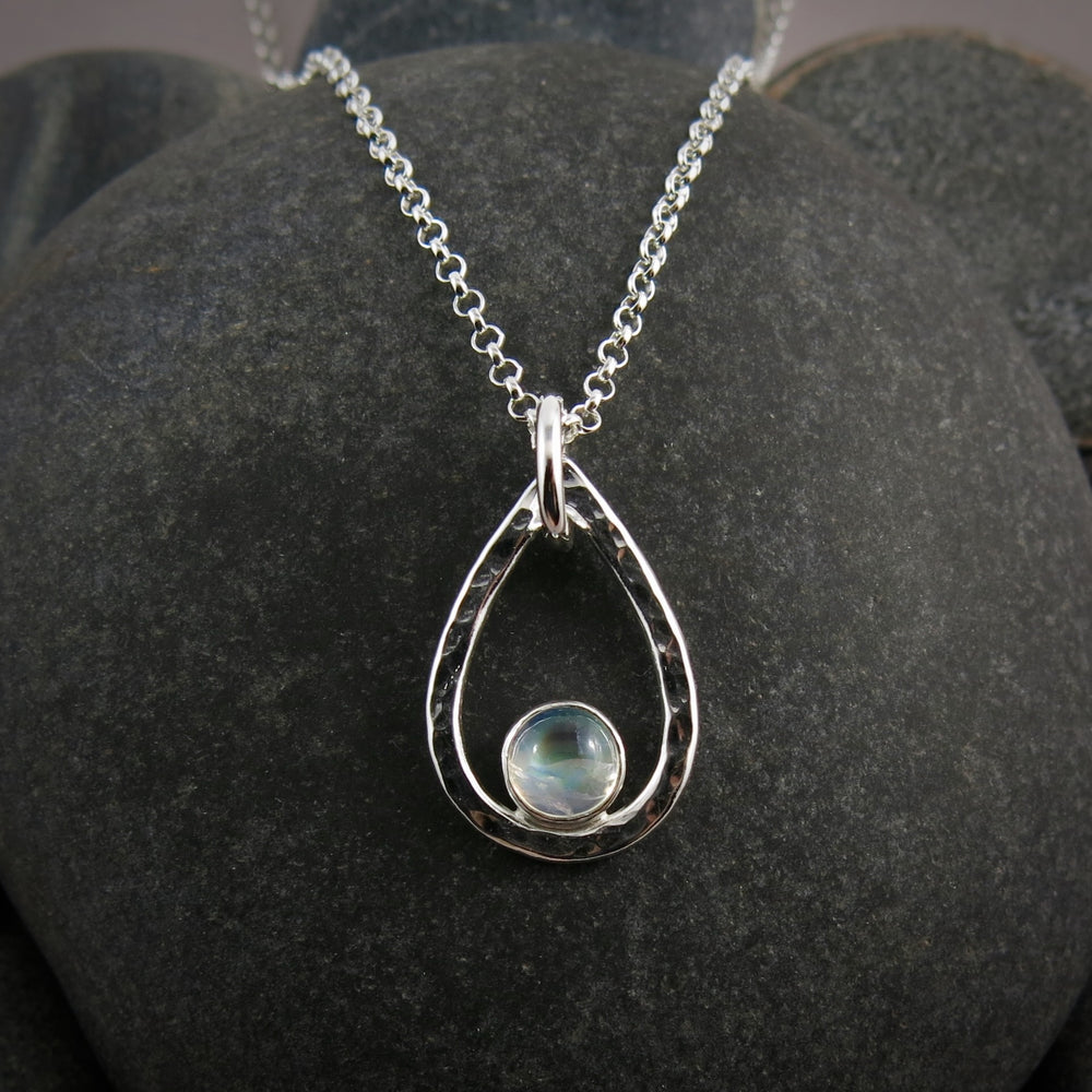 A teardrop shaped necklace is displayed on a black beach rock.  The hammer textured drop is accented by a round rainbow moonstone that sits in the curve at the bottom of the pendant.  A silver rolo chain completes the piece.