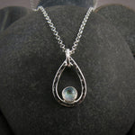 A teardrop shaped necklace is displayed on a black beach rock.  The hammer textured drop is accented by a round rainbow moonstone that sits in the curve at the bottom of the pendant.  A silver rolo chain completes the piece.