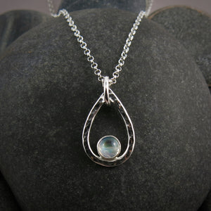 A teardrop shaped necklace is displayed on a black beach rock.  The hammer textured drop is accented by a round rainbow moonstone that sits in the curve at the bottom of the pendant.  A silver rolo chain completes the piece.