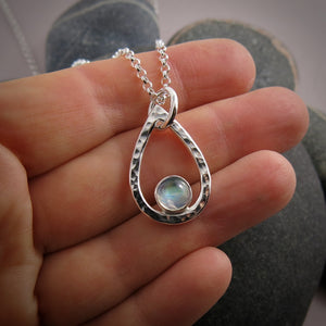 A teardrop shaped necklace is displayed on a hand.  The hammer textured drop is accented by a round rainbow moonstone that sits in the curve at the bottom of the pendant.  A silver rolo chain completes the piece.