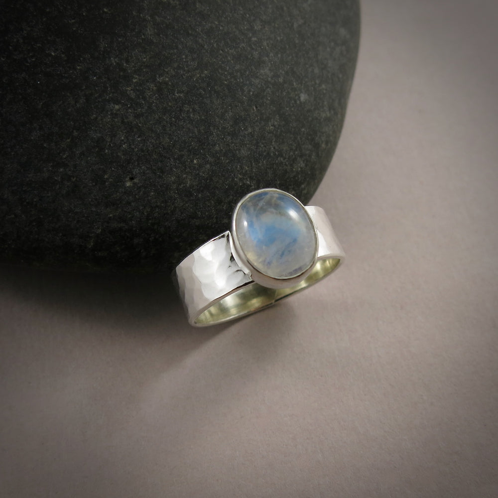 A silver and gemstone ring is displayed on a gray background.  The hammer textured ring features an oval rainbow moonstone on top.  