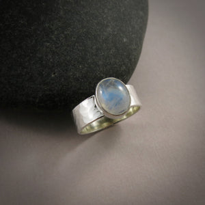 A silver and gemstone ring is displayed on a gray background.  The hammer textured ring features an oval rainbow moonstone on top.  