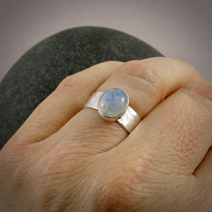 A silver and gemstone ring is displayed on a finger.  The hammer textured ring features an oval rainbow moonstone on top.  