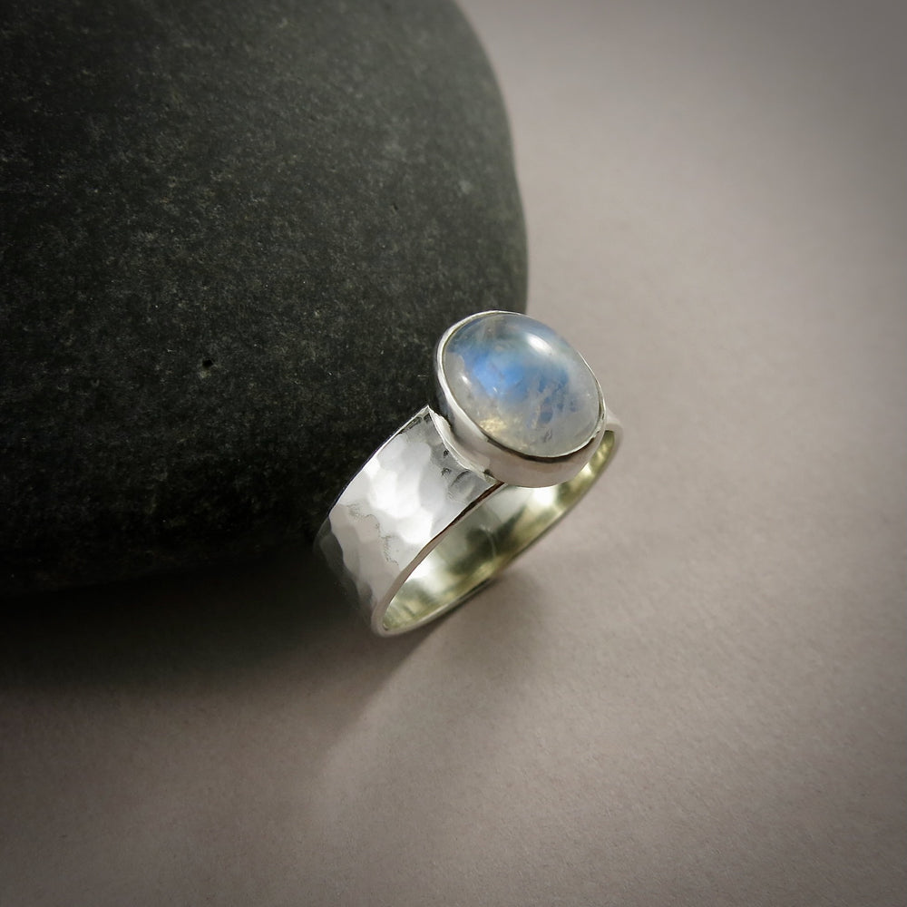 A silver and gemstone ring is displayed on a gray background.  The hammer textured ring features an oval rainbow moonstone on top.  