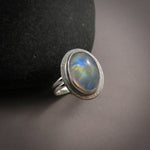 A silver ring with a rainbow moonstone is displayed on a dark surface.  The oval moonstone shows a variety of colours from orange and yellow to green and blue.
