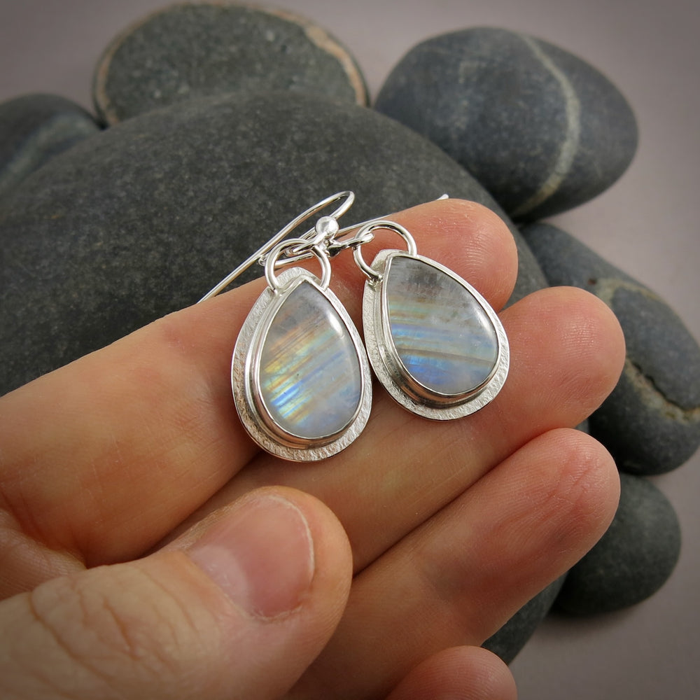 Teardrop-shaped silver earrings with rainbow moonstone stones held between fingers against a stone background.