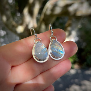Teardrop-shaped silver earrings with rainbow moonstone stones held between fingers against a natural background.