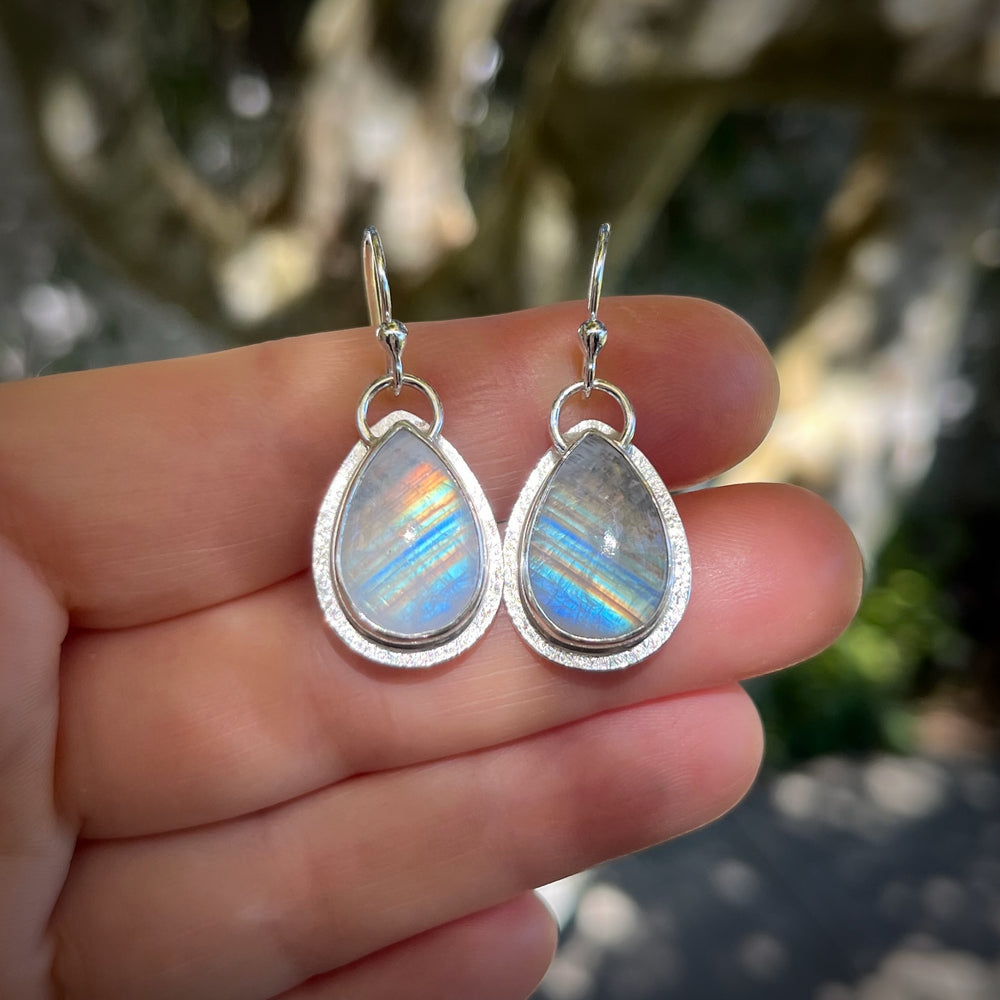 Teardrop-shaped silver earrings with rainbow moonstone stones held between fingers against a natural background.