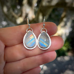 Teardrop-shaped silver earrings with rainbow moonstone stones held between fingers against a natural background.