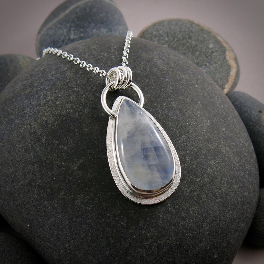 A moonstone pendant is displayed on a black beach rock.  The teardrop shaped moonstone is white with an inner blue glow.  The stone is bezel set in a silver setting and hangs from a silver rolo chain.