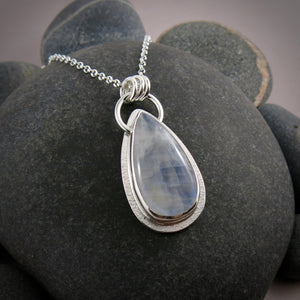 A moonstone pendant is displayed on a black beach rock.  The teardrop shaped moonstone is white with an inner blue glow.  The stone is bezel set in a silver setting and hangs from a silver rolo chain.