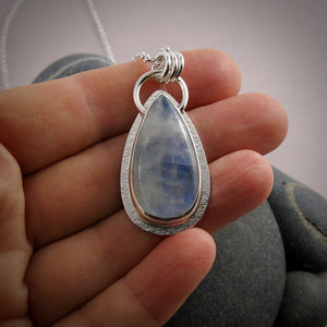 A moonstone pendant is displayed on a hand.  The teardrop shaped moonstone is white with an inner blue glow.  The stone is bezel set in a silver setting and hangs from a silver rolo chain.