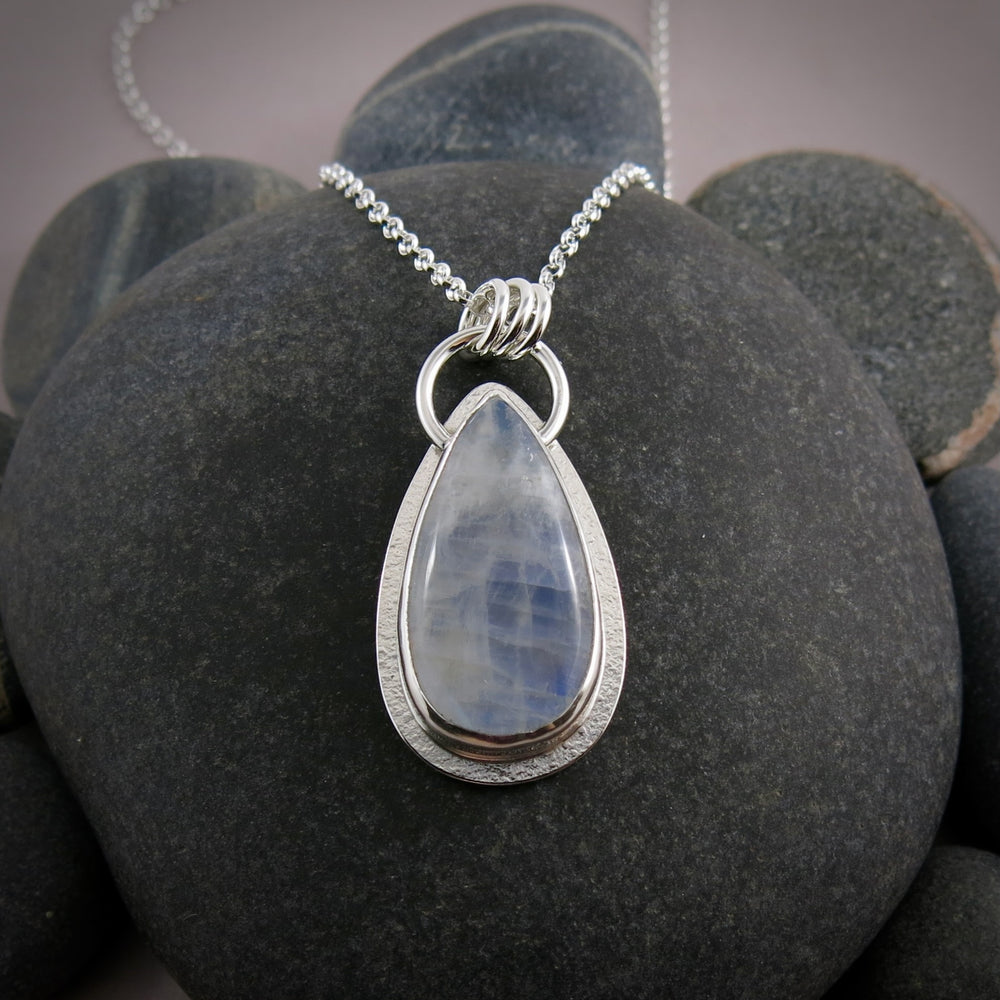 A moonstone pendant is displayed on a black beach rock.  The teardrop shaped moonstone is white with an inner blue glow.  The stone is bezel set in a silver setting and hangs from a silver rolo chain.