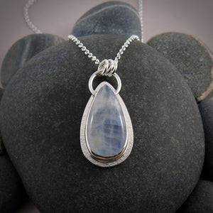 A moonstone pendant is displayed on a black beach rock.  The teardrop shaped moonstone is white with an inner blue glow.  The stone is bezel set in a silver setting and hangs from a silver rolo chain.