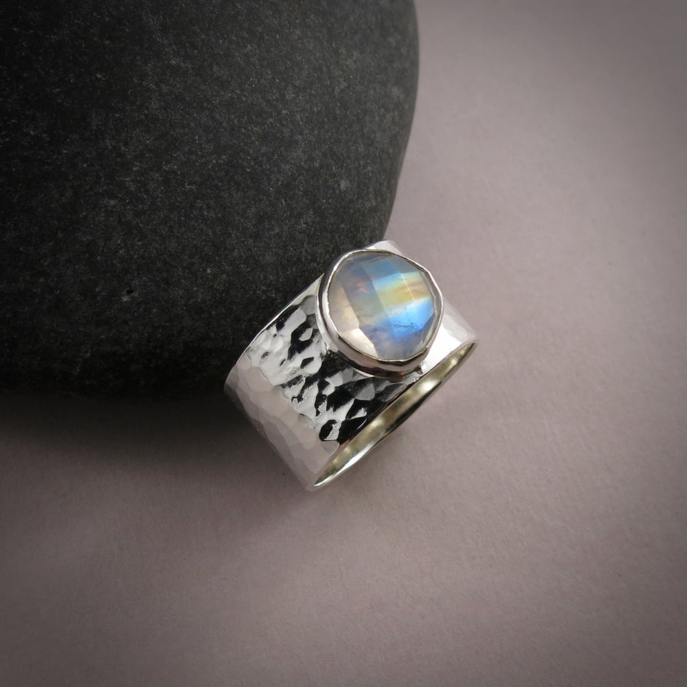 A wide, hammer textured silver ring is displayed on a gray background.  The ring features a bezel set stone on top.  The gem is a rose-cut rainbow moonstone displaying a lovely flash of orange, gold and blue.