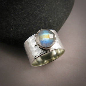 A wide, hammer textured silver ring is displayed on a gray background.  The ring features a bezel set stone on top.  The gem is a rose-cut rainbow moonstone displaying a lovely flash of orange, gold and blue.