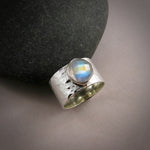 A wide, hammer textured silver ring is displayed on a gray background.  The ring features a bezel set stone on top.  The gem is a rose-cut rainbow moonstone displaying a lovely flash of orange, gold and blue.