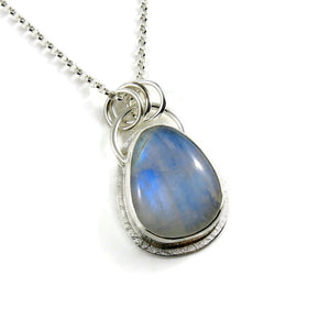 A silver necklace with a gemstone pendant is displayed on a white background.  The pendant features a rounded teardrop shaped rainbow moonstone with blue flash.  The gemstone is set in a silver bezel and a subtly textured silver "halo" surround.