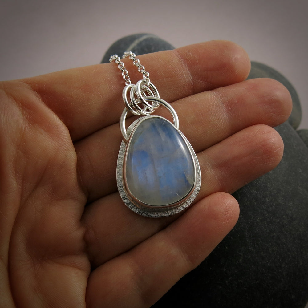 A silver necklace with a gemstone pendant is displayed on a hand.  The pendant features a rounded teardrop shaped rainbow moonstone with blue flash.  The gemstone is set in a silver bezel and a subtly textured silver "halo" surround.
