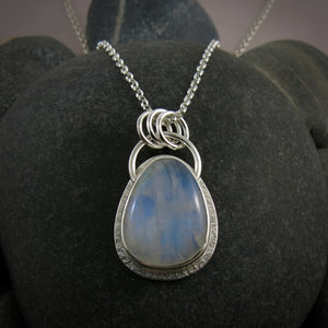 A silver necklace with a gemstone pendant is displayed on a black beach rock.  The pendant features a rounded teardrop shaped rainbow moonstone with blue flash.  The gemstone is set in a silver bezel and a subtly textured silver "halo" surround.