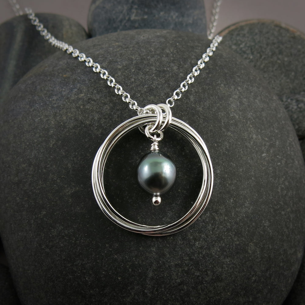 A silver knot pendant with a lustrous grey-green Tahitian pearl suspended within the knot is displayed against a black beach rock.