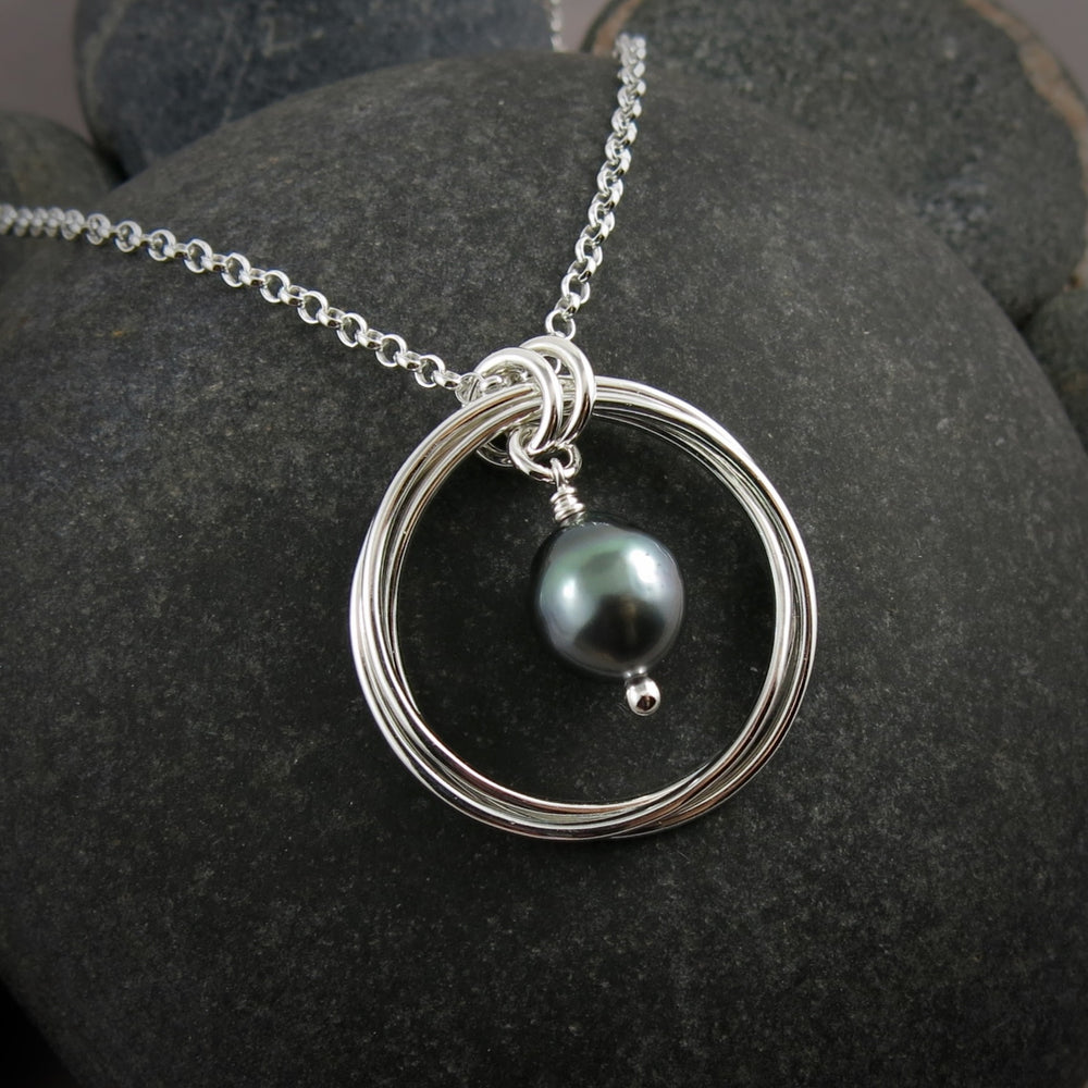 A silver knot pendant with a lustrous grey-green Tahitian pearl suspended within the knot is displayed against a black beach rock.