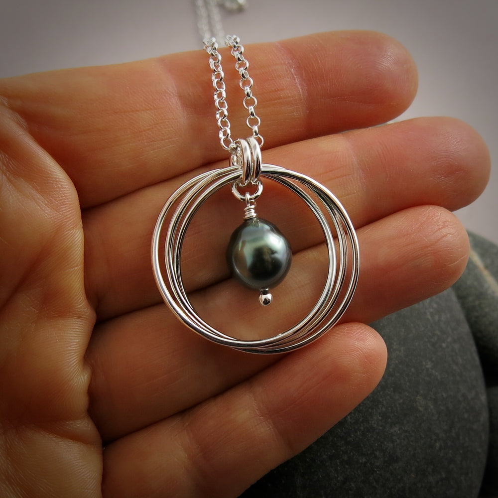 A silver knot pendant with a lustrous grey-green Tahitian pearl suspended within the knot is displayed against a hand.