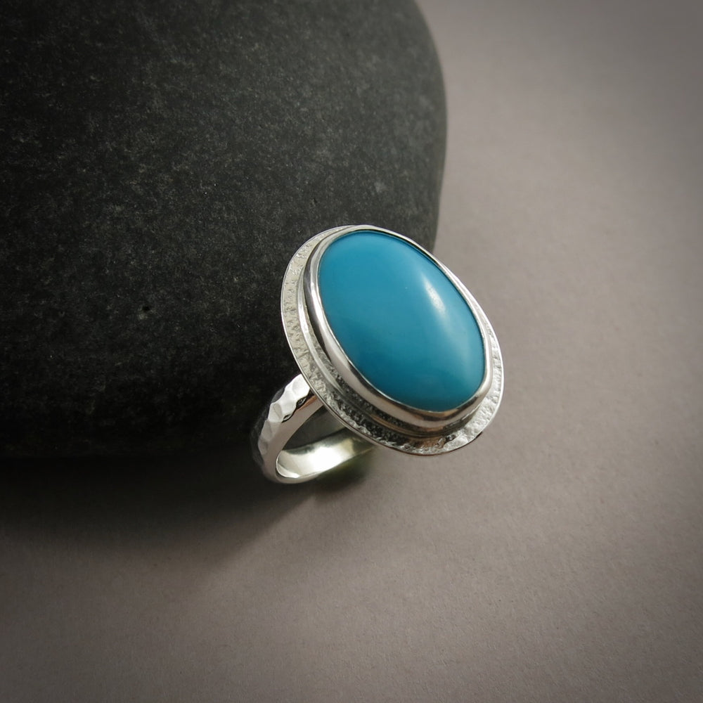 A sterling silver ring with a bright blue oval turquoise gemstone is displayed on a dark background against a black beach rock.  The ring has a hammer textured band and a subtly textured "halo" surrounding the turquoise.