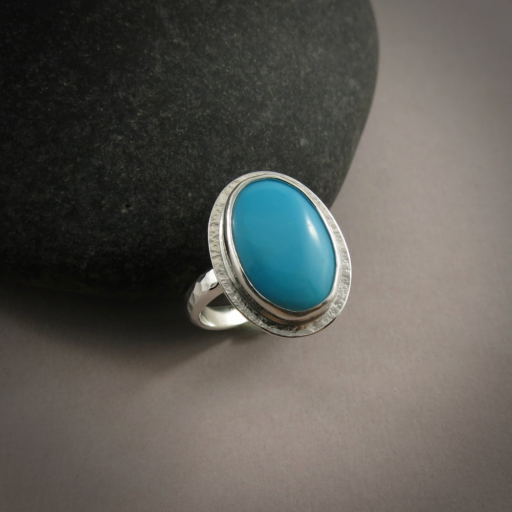 A sterling silver ring with a bright blue oval turquoise gemstone is displayed on a dark background against a black beach rock.  The ring has a hammer textured band and a subtly textured "halo" surrounding the turquoise.