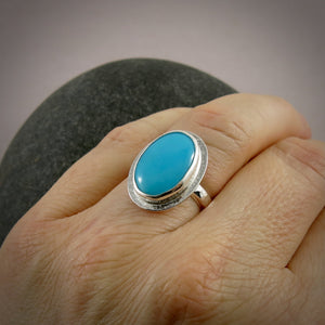 A sterling silver ring with a bright blue oval turquoise gemstone is displayed on a hand.  The ring has a hammer textured band and a subtly textured "halo" surrounding the turquoise.
