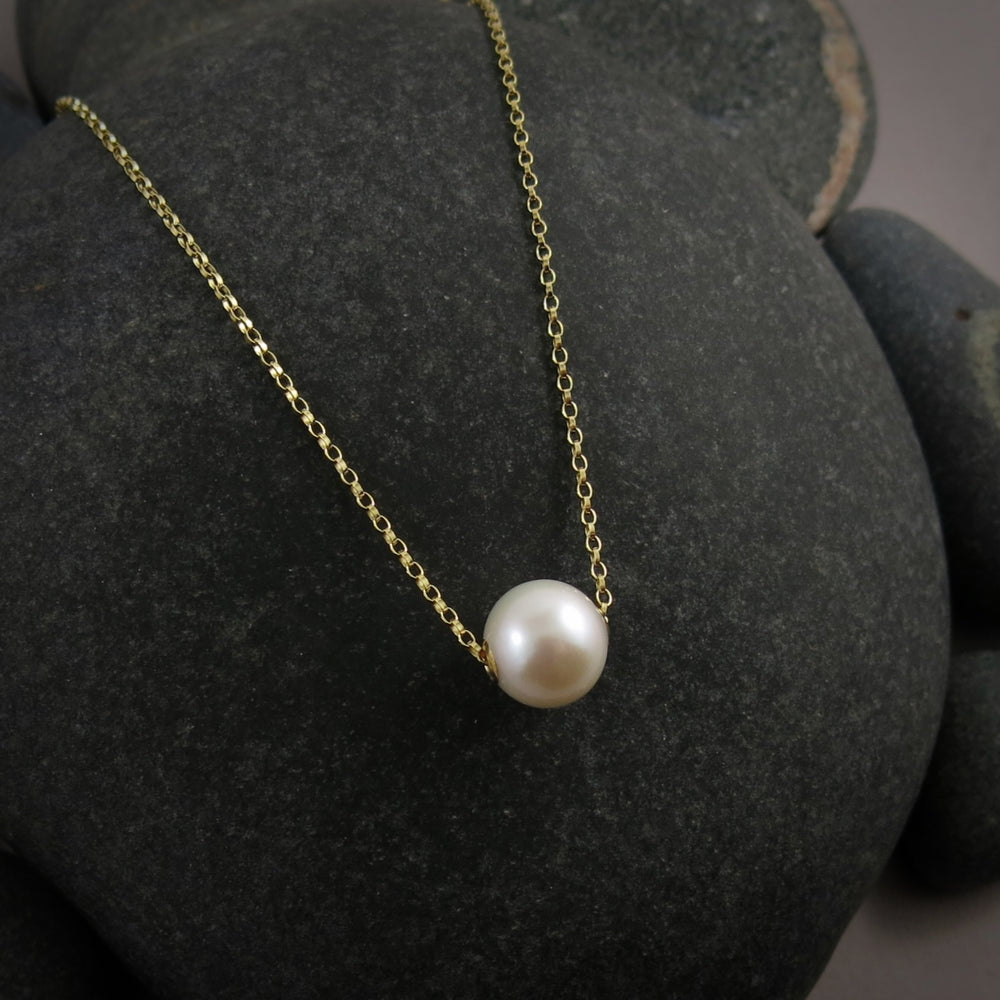A single pearl necklace on a gold chain is displayed on a black beach rock.  The round white pearl can slide back and forth on the flat gold rolo chain that goes through the middle of the pearl.