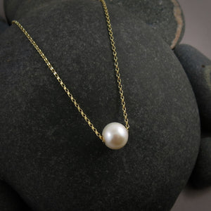 A single pearl necklace on a gold chain is displayed on a black beach rock.  The round white pearl can slide back and forth on the flat gold rolo chain that goes through the middle of the pearl.