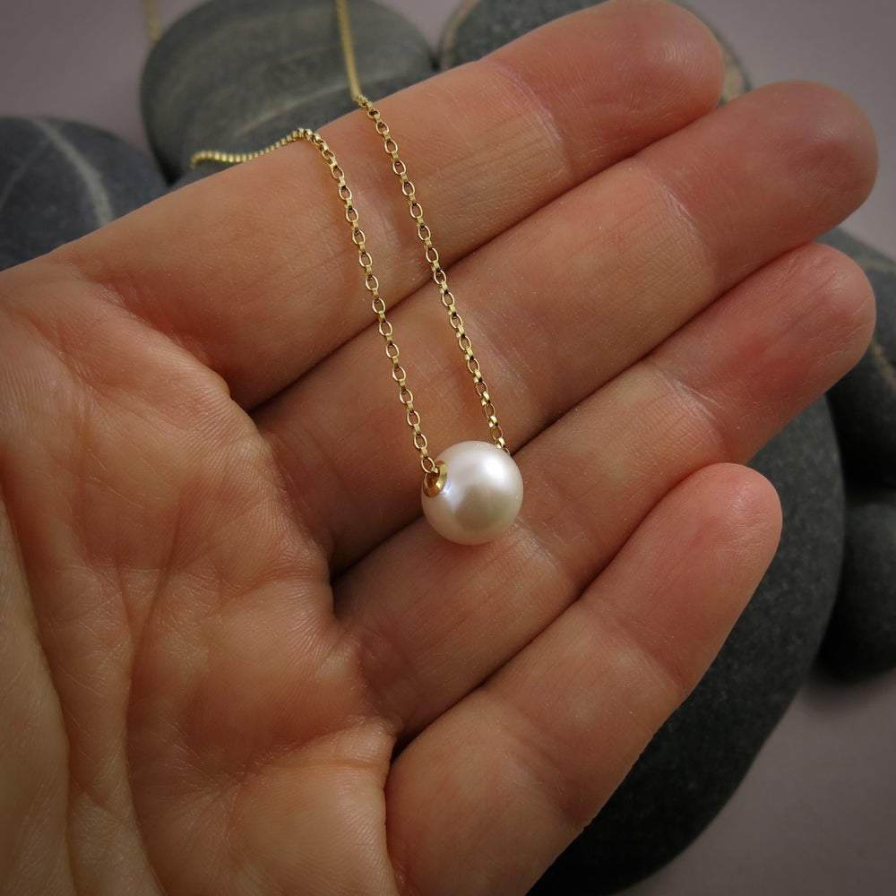 A single pearl necklace on a gold chain is displayed on a hand for size reference.  The round white pearl can slide back and forth on the flat gold rolo chain that goes through the middle of the pearl.