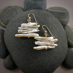 A pair of white pearl earrings are displayed on a black beach rock.  The earrings feature a stack of five organically shaped keshi toothpick earrings on a gold headpin with gold ear wires.