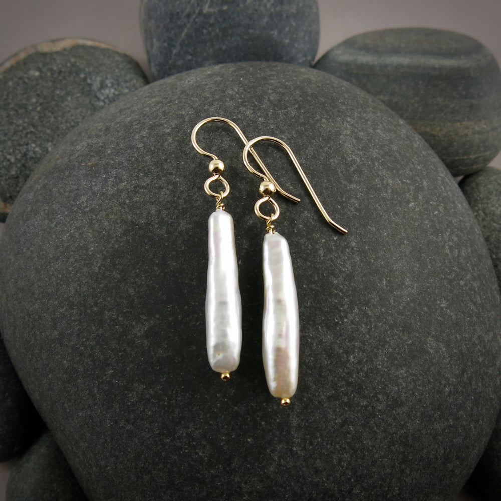 A pair of pearl earrings are displayed on a black beach rock.  The rectangular white stick pearls hang from gold filled ear wires.