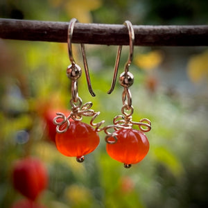 A pair of earrings are hanging on a branch with blurred natural background.  The earrings feature carved orange carnelian beads that look like tiny pumpkins with curled gold filled wire that looks like vines atop.  They hang from gold filled ear wires.